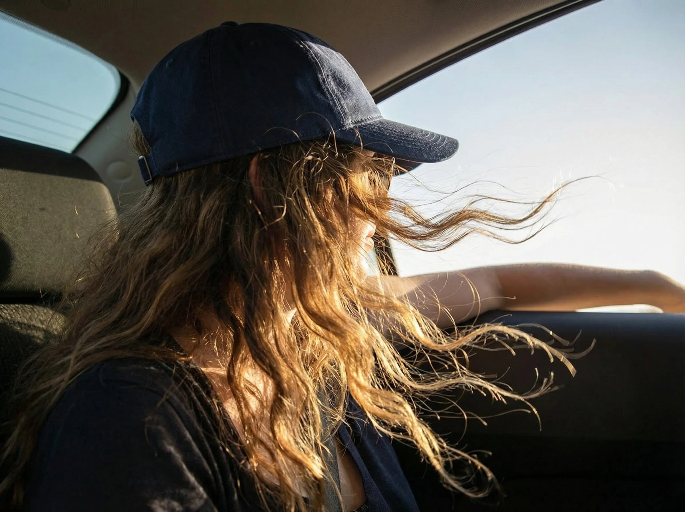 A woman with long, curly hair wearing a baseball cap, sitting in the backseat of a car, looking out the window with her arm resting on the door.