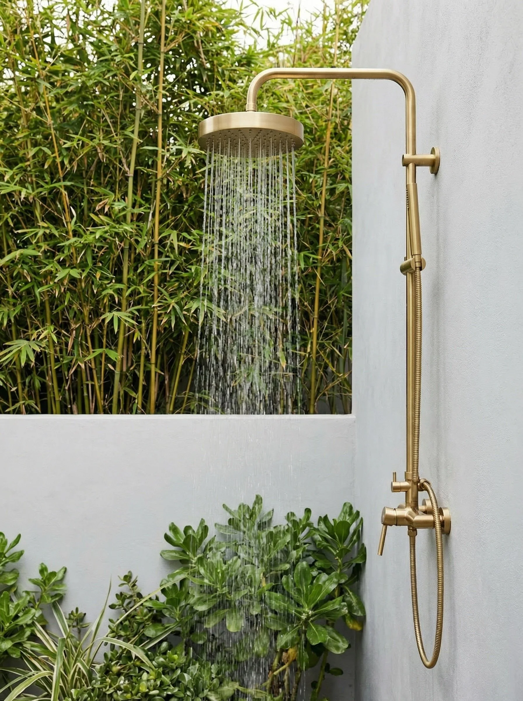 Outdoor shower with gold fixtures mounted on a gray wall, with greenery in the background and foreground.