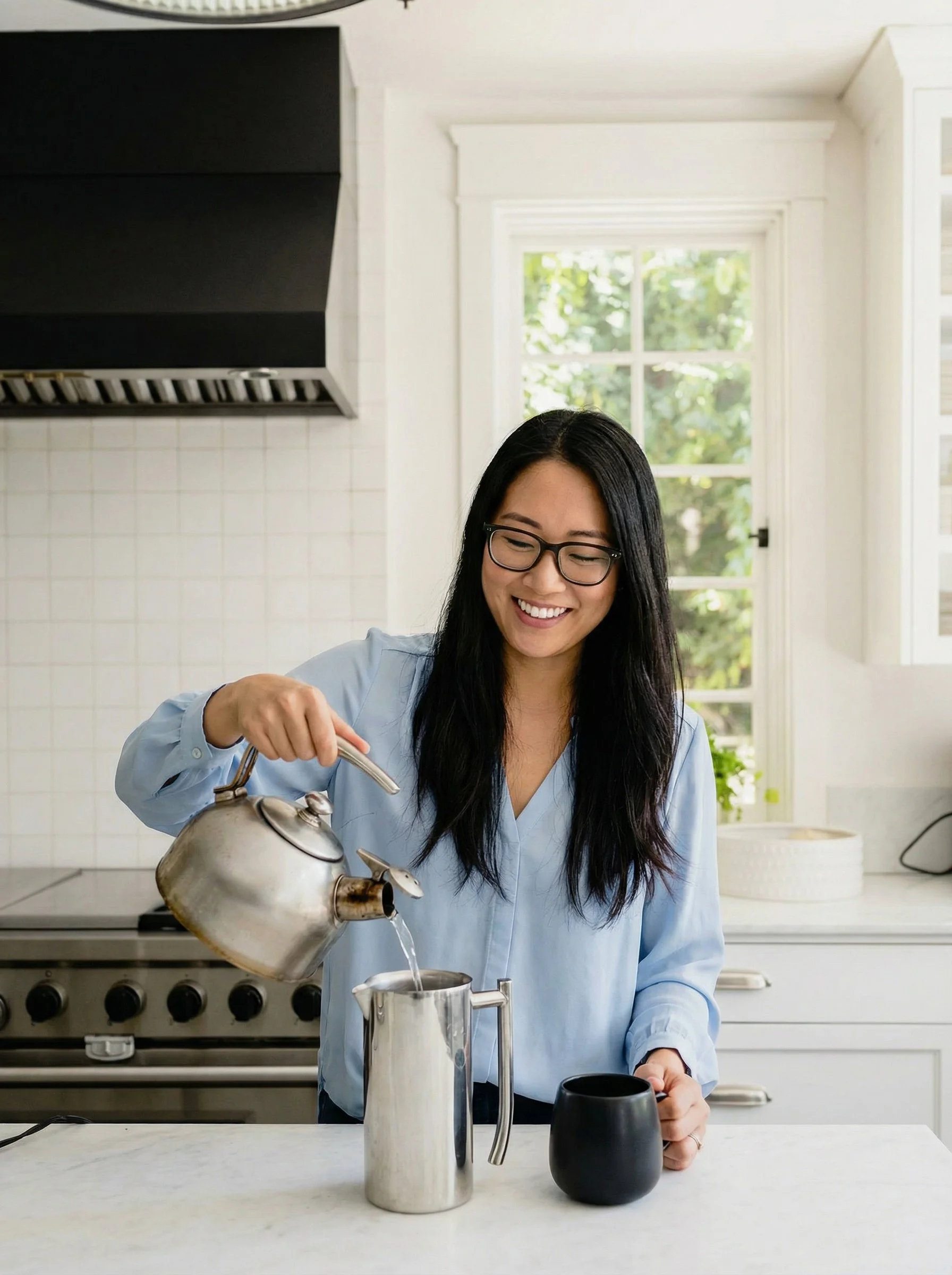A woman with glasses and long black hair smiling and pouring hot water from a metal kettle into a French press on a white kitchen countertop with a black mug nearby.