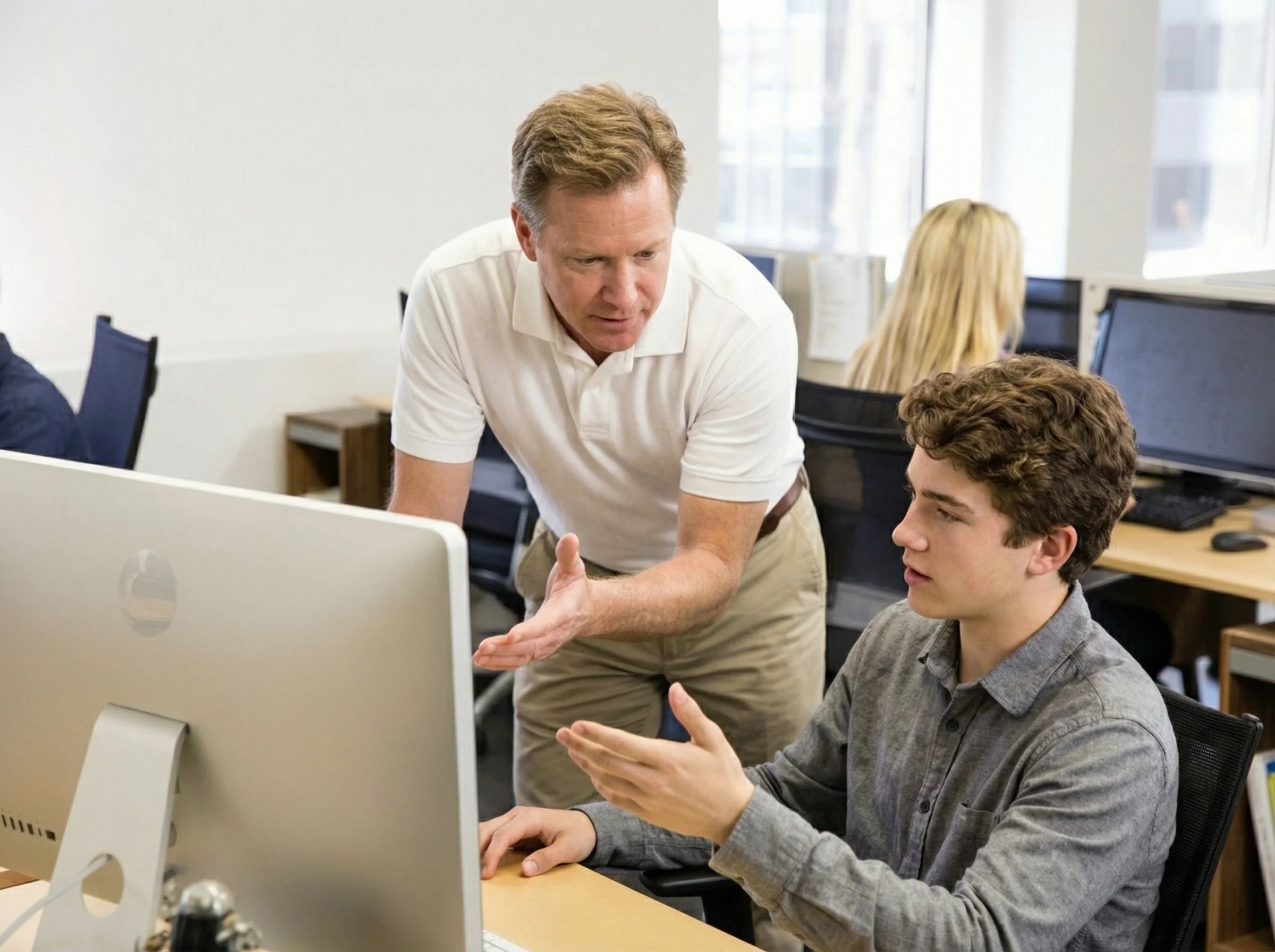 An older man in a white polo shirt is standing and talking to a young man with curly hair, who is sitting at a desk in front of a desktop computer. They appear to be having a discussion or explaining something. In the background, there are other peop
