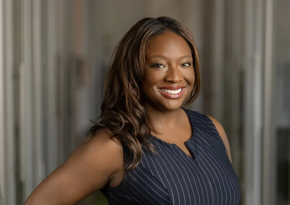 A woman with shoulder-length brown hair, smiling, wearing a navy blue sleeveless top with thin white stripes, standing in front of a blurred background.