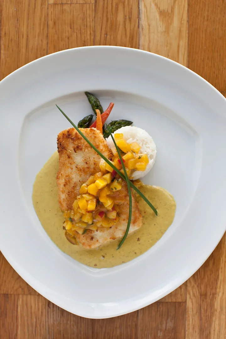 Breaded fish fillet topped with yellow corn salsa, served with hush puppy-mashed potato ball, green beans, and a light yellow sauce on a white plate
