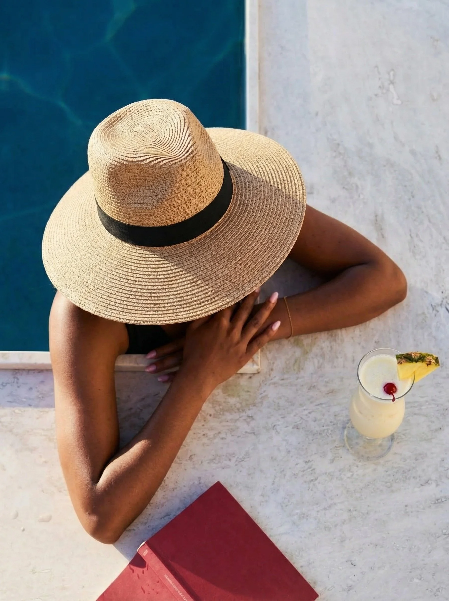 A person wearing a wide-brimmed straw hat with a black band sitting at a marble table by a swimming pool, with a tropical drink garnished with pineapple and cherry.