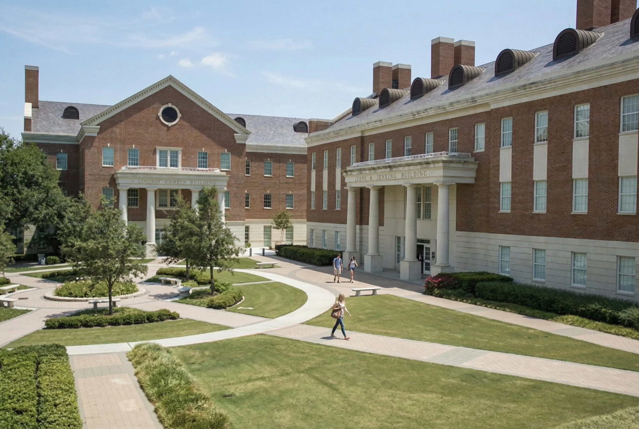 A college campus with a large brick building featuring white columns and multiple windows. The building has signs indicating it is the Lindsay Carey Building and the Stone & Jexing Building. There are well-maintained lawns, trees, pathways, and a few