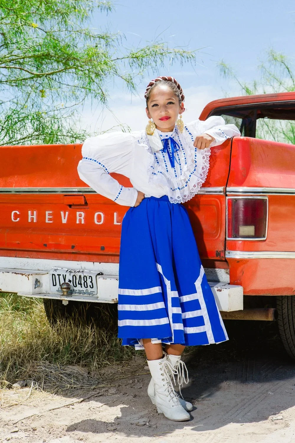 A young girl in traditional Mexican dress standing in front of a red Chevrolet pickup truck outdoors on a sunny day.