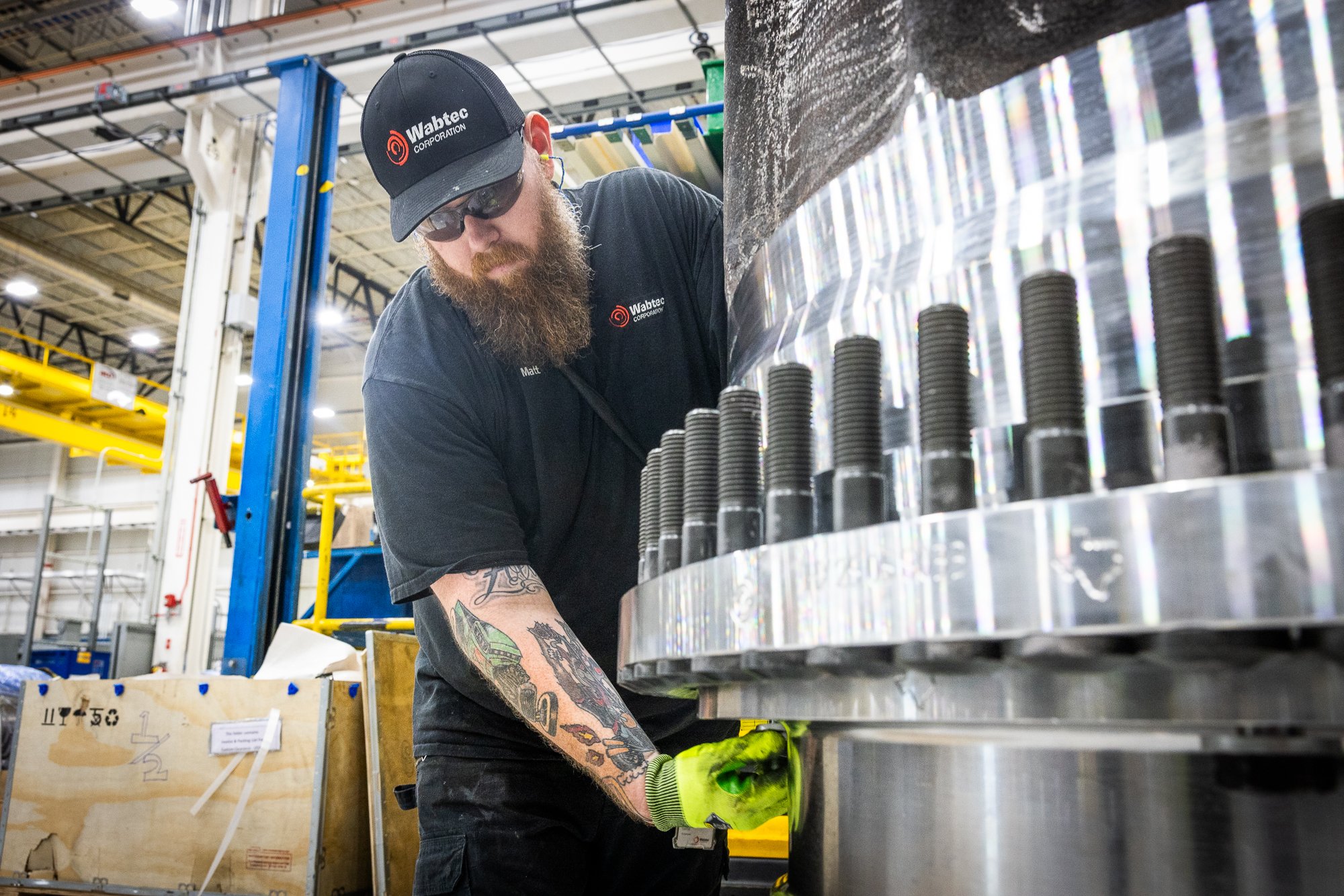 A man with a beard, tattoos on his arm, wearing a black Wabtec Corporation cap, sunglasses, and gloves is working on a large metal component with bolts in an industrial manufacturing setting.