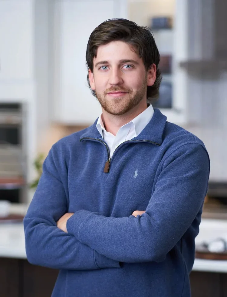 Young man with brown hair and beard, wearing a blue quarter-zip sweater over a white shirt, standing with arms crossed in a modern kitchen.