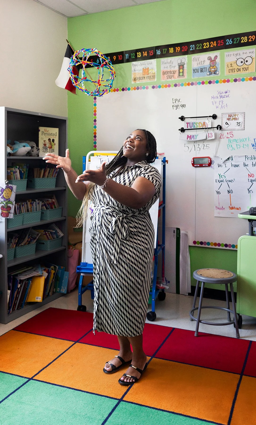 Woman in a striped dress with braids standing in a colorful classroom, playing with a hanging globe-shaped structure, surrounded by educational posters and books.