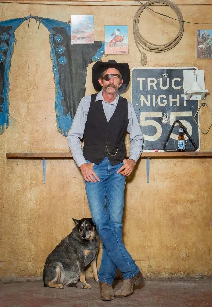 A man with a mustache and an eyepatch dressed in a cowboy hat, vest, and jeans, standing with one foot on the ground and the other crossed over, next to a dog sitting against a rustic wall. The setting appears to be a Western-themed bar or saloon, wi