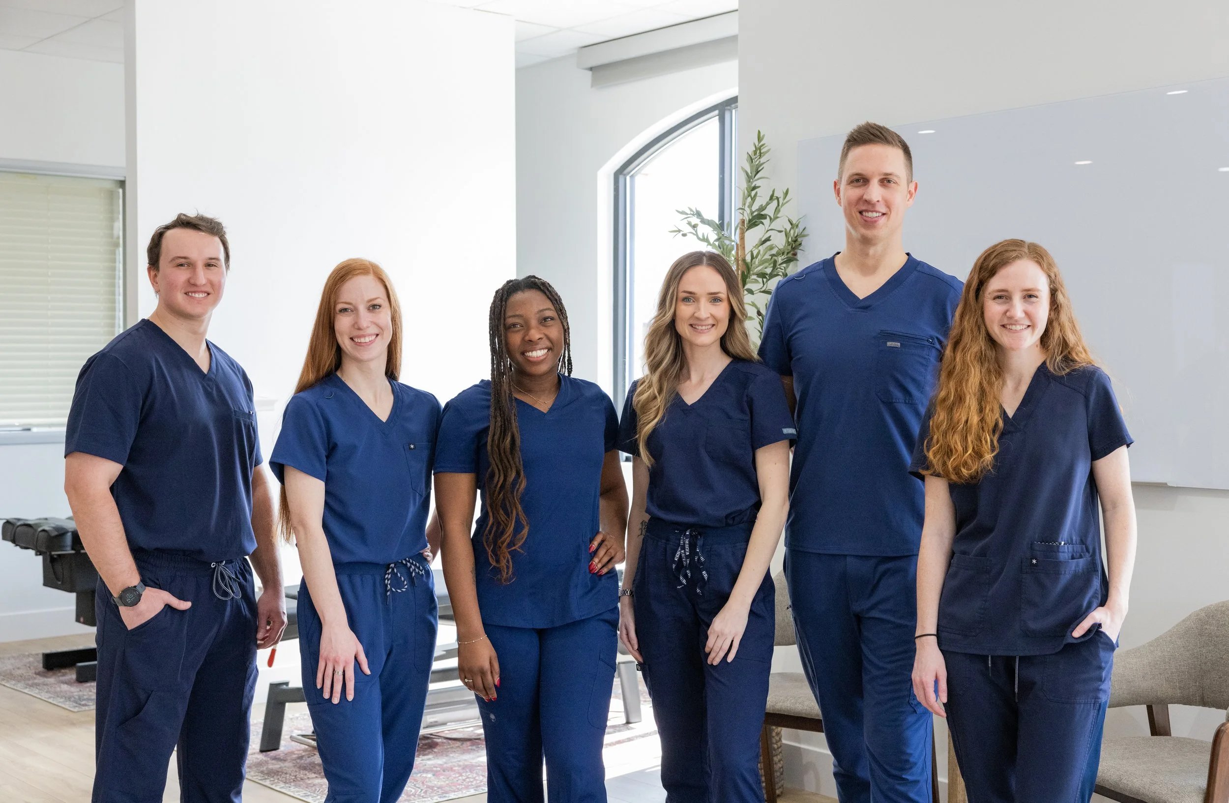 Group of six healthcare professionals wearing navy scrubs standing together in a well-lit room, smiling at the camera.