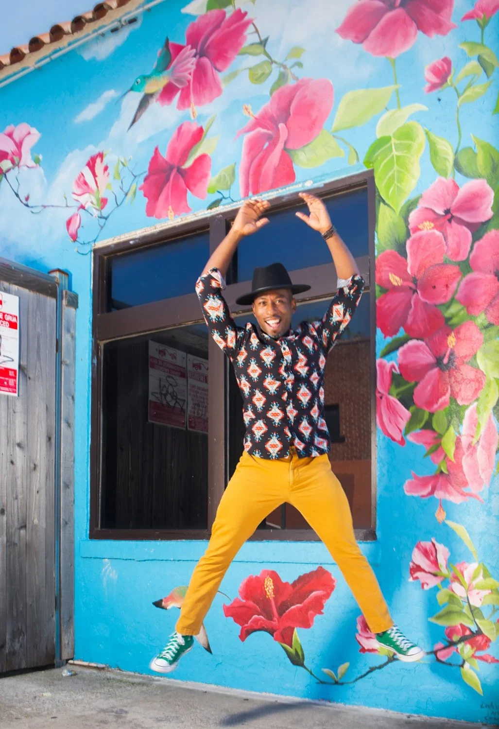A man wearing a black hat, colorful patterned shirt, mustard pants, and green sneakers jumping in front of a vibrant painted wall with pink flowers, green leaves, blue sky, and a hummingbird mural.