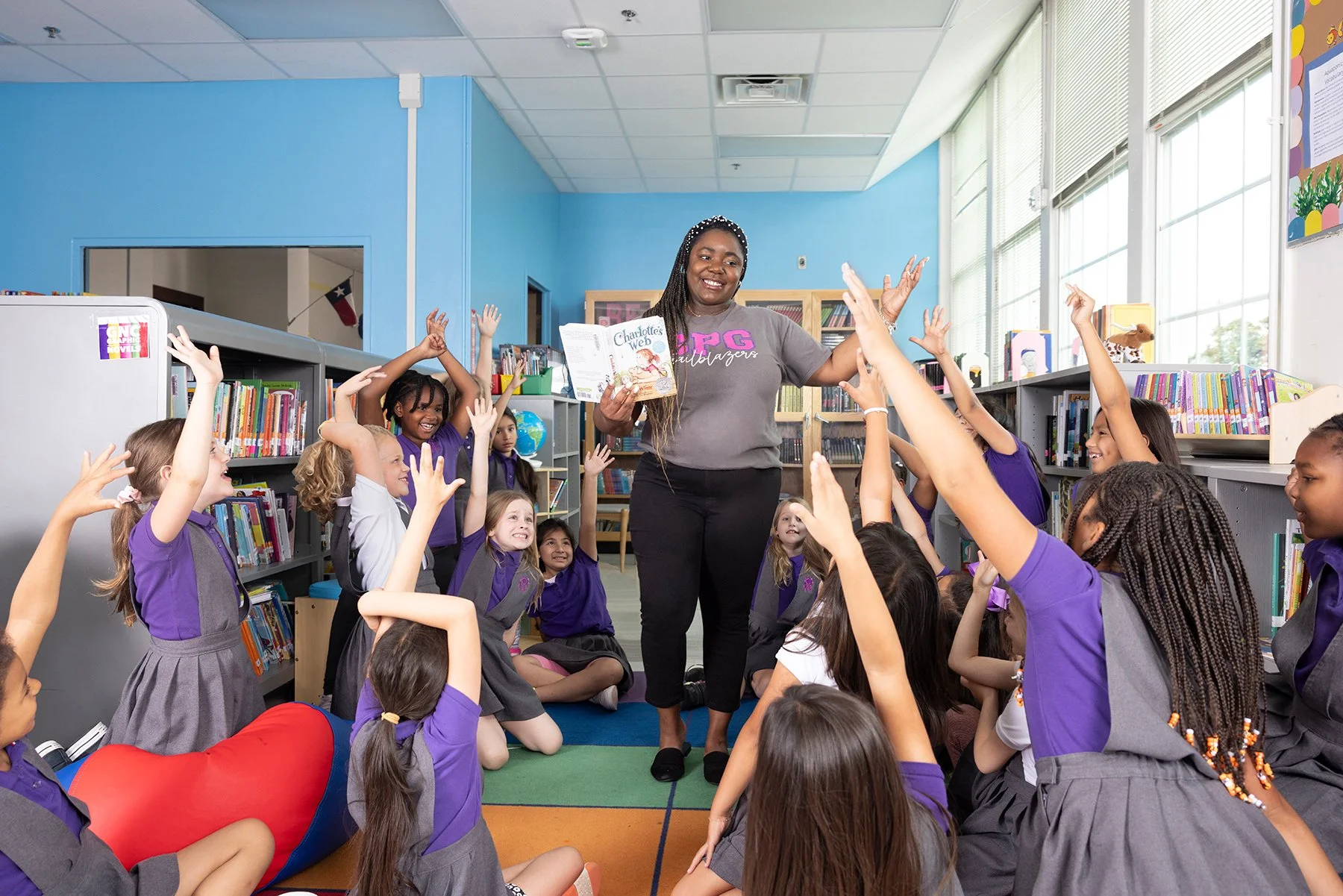 A classroom with a teacher reading a book to a group of children, who are sitting cross-legged on colorful carpet squares with their hands raised eagerly.