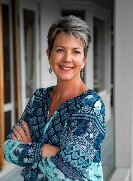 A smiling middle-aged woman with short gray hair, wearing a blue patterned blouse, standing on a porch with windows in the background.