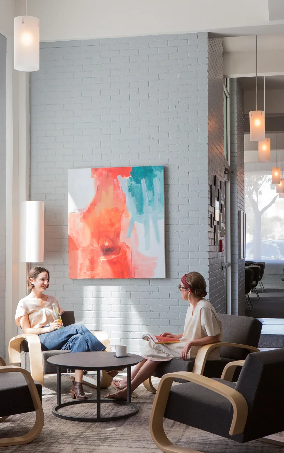 Two women are sitting and talking in a modern, well-lit waiting room with gray chairs, a black coffee table, and white brick walls decorated with a piece of abstract art. One woman holds a beverage, and the other has a book.