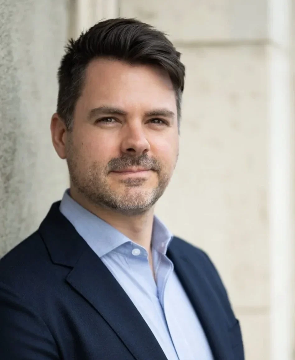 A man in a dark suit and light blue shirt standing outdoors near a wall, looking at the camera.