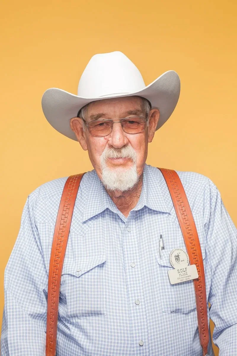 An elderly man wearing a white cowboy hat, glasses, with a beard and mustache, dressed in a light blue checkered shirt with suspenders, standing against a yellow background.