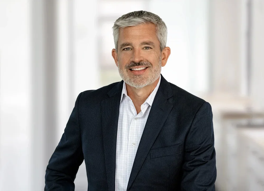 A middle-aged man with gray hair and a beard, smiling, wearing a dark blazer and a white dress shirt, in an office setting with soft lighting and blurred background.