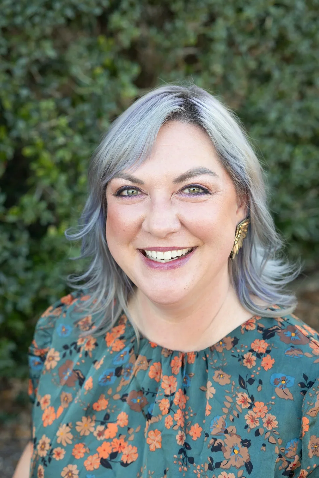 A woman with light gray hair colored with blue highlights, smiling outdoors against greenery, wearing a floral top and gold earrings.