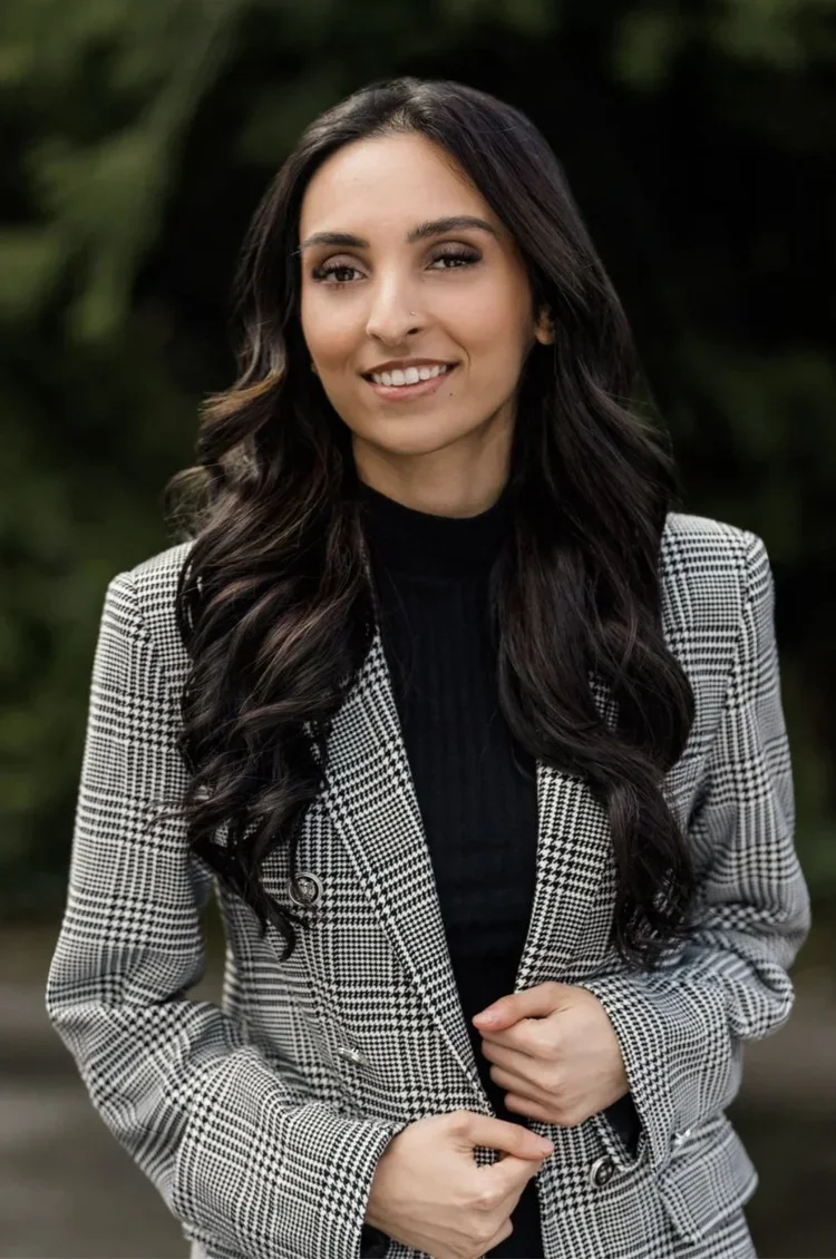 Young woman outdoors smiling, wearing a black top and plaid blazer with long dark hair.
