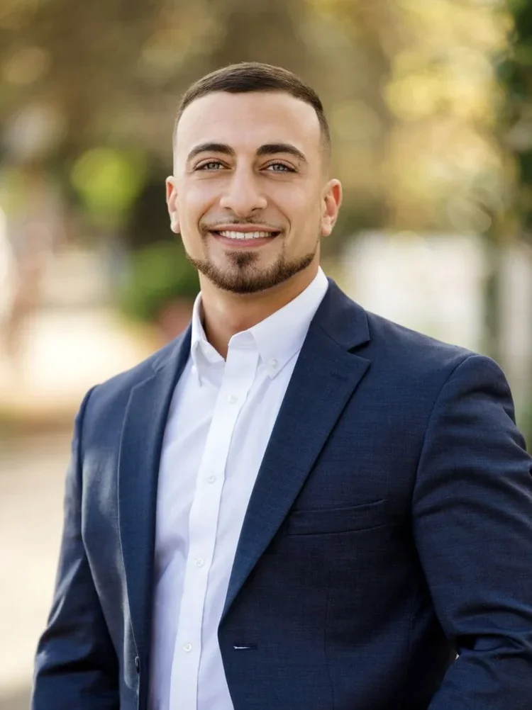A smiling man in a navy blue suit and white dress shirt standing outdoors with blurred trees in the background