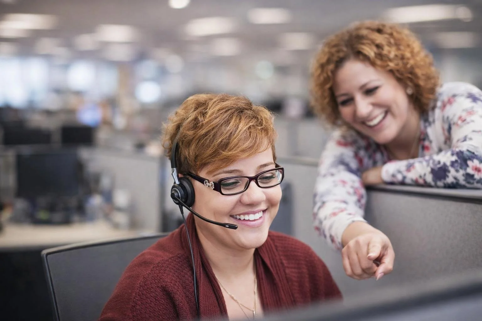 Two women working together in an office, one wearing a headset and glasses, smiling at her computer, while the other woman leans over the cubicle, smiling and pointing at the screen.