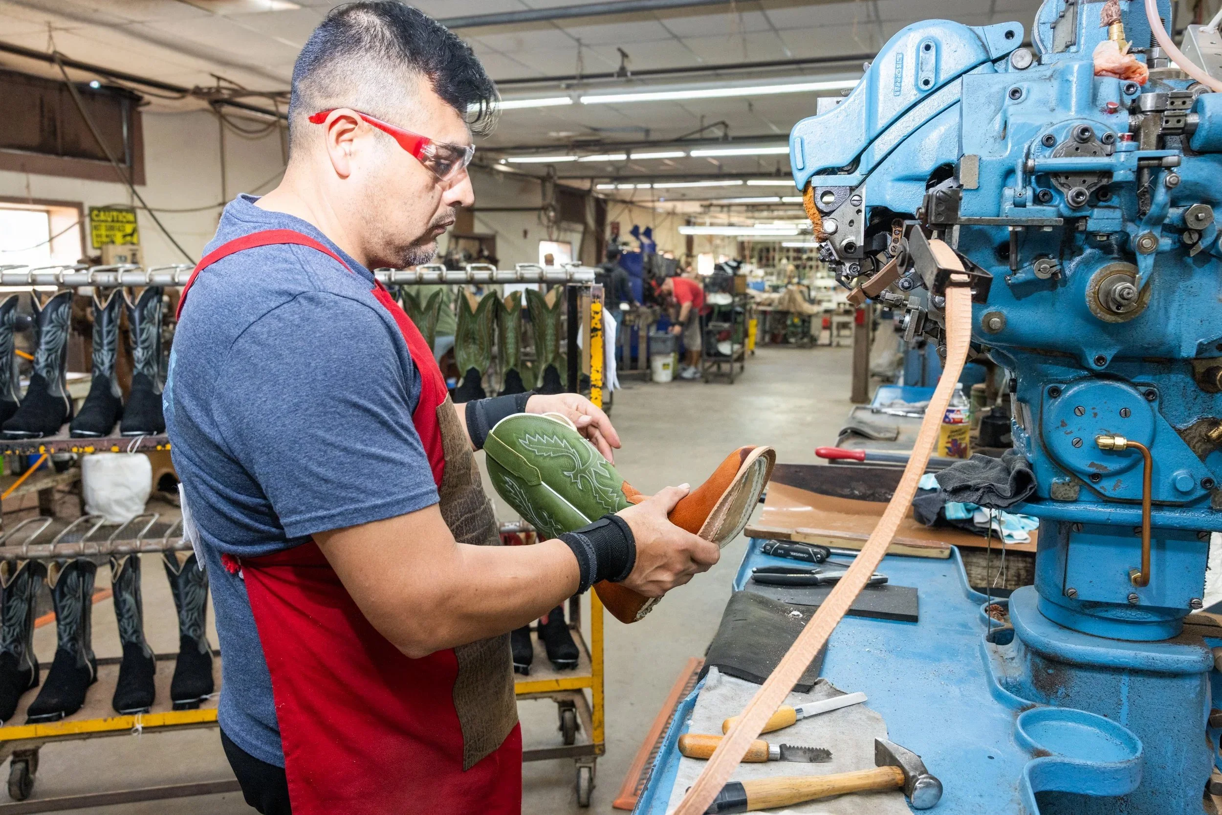 A worker wearing safety glasses and a red apron working on a leather shoe in a shoe manufacturing factory.