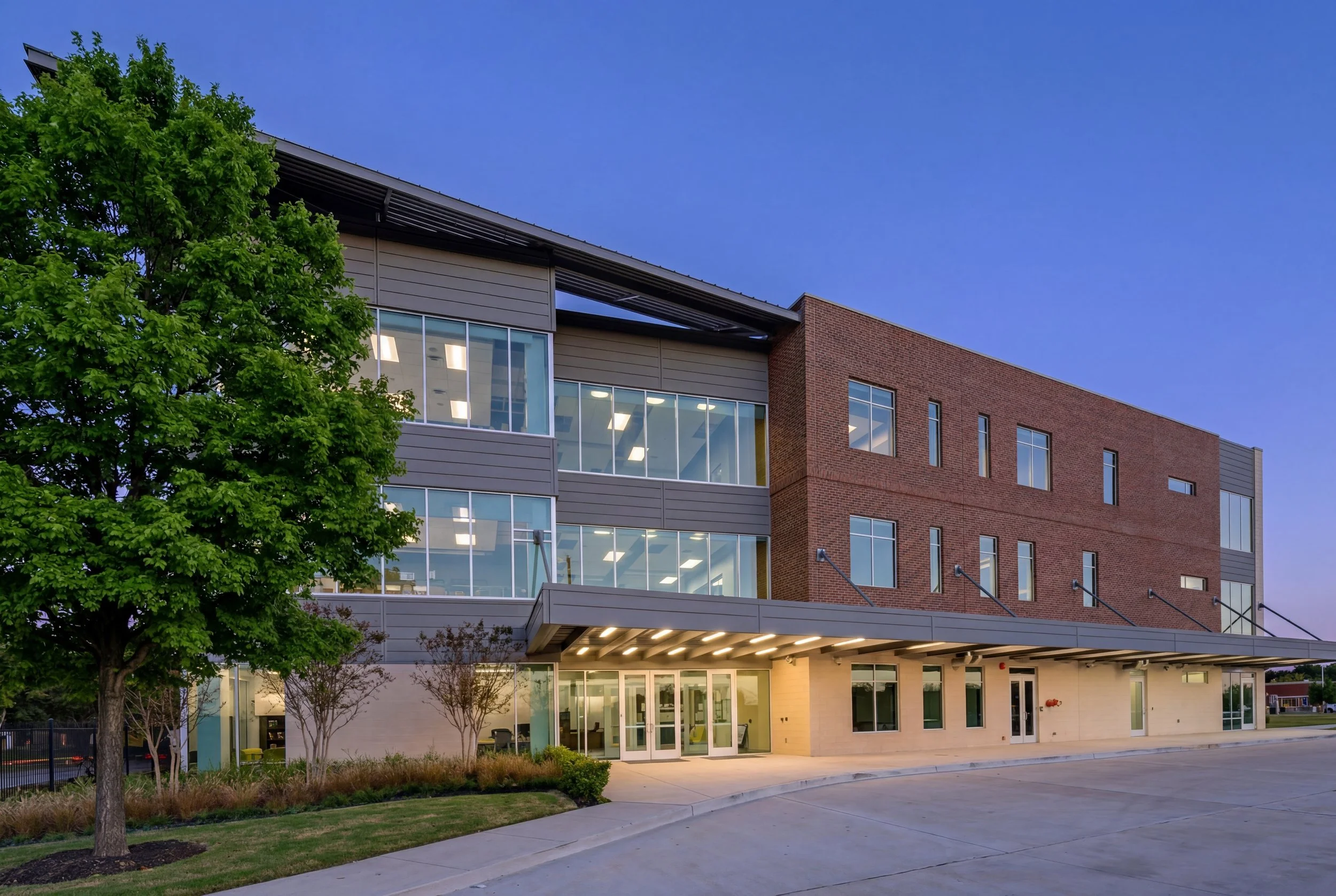 Exterior view of a modern multi-story building with glass windows, brick and gray siding, surrounded by trees, under a clear blue sky at dusk.