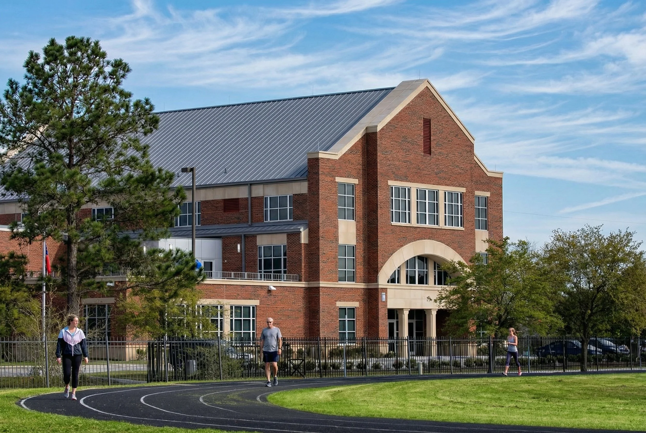 A brick school building with large windows and a metal roof, located behind a black metal fence. There is a grassy field with a running track in the foreground, and three people walking on or near the track. Trees are around the building, and a partl