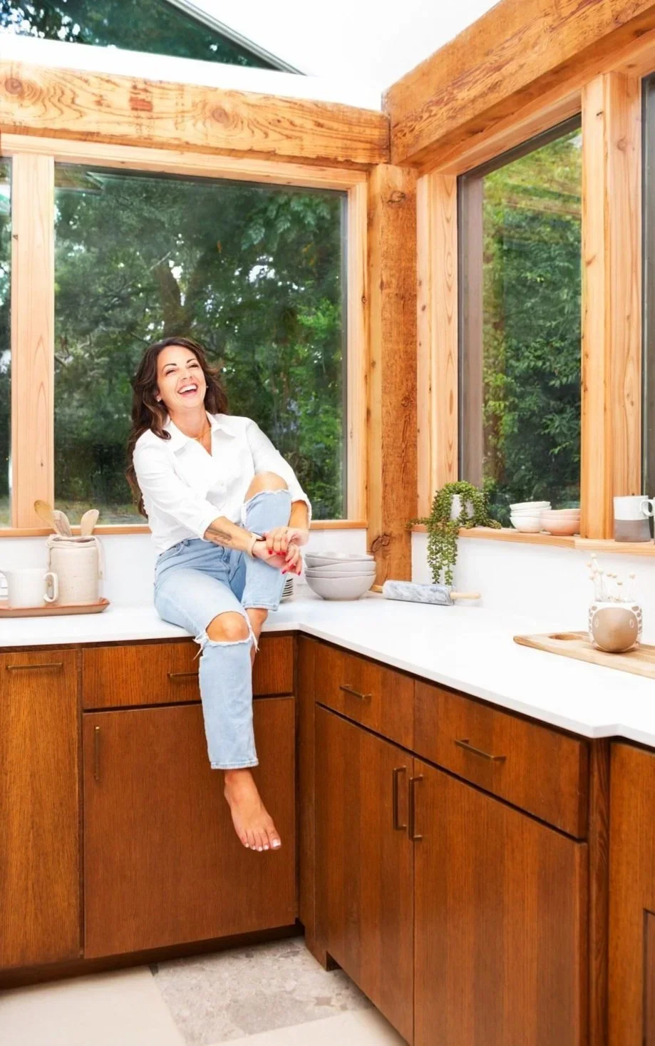 A woman with long brown hair, wearing a white shirt and ripped jeans, sitting on the kitchen counter with one leg crossed over the other, smiling and laughing inside a cozy kitchen with wooden cabinets and large windows showing green trees outside.