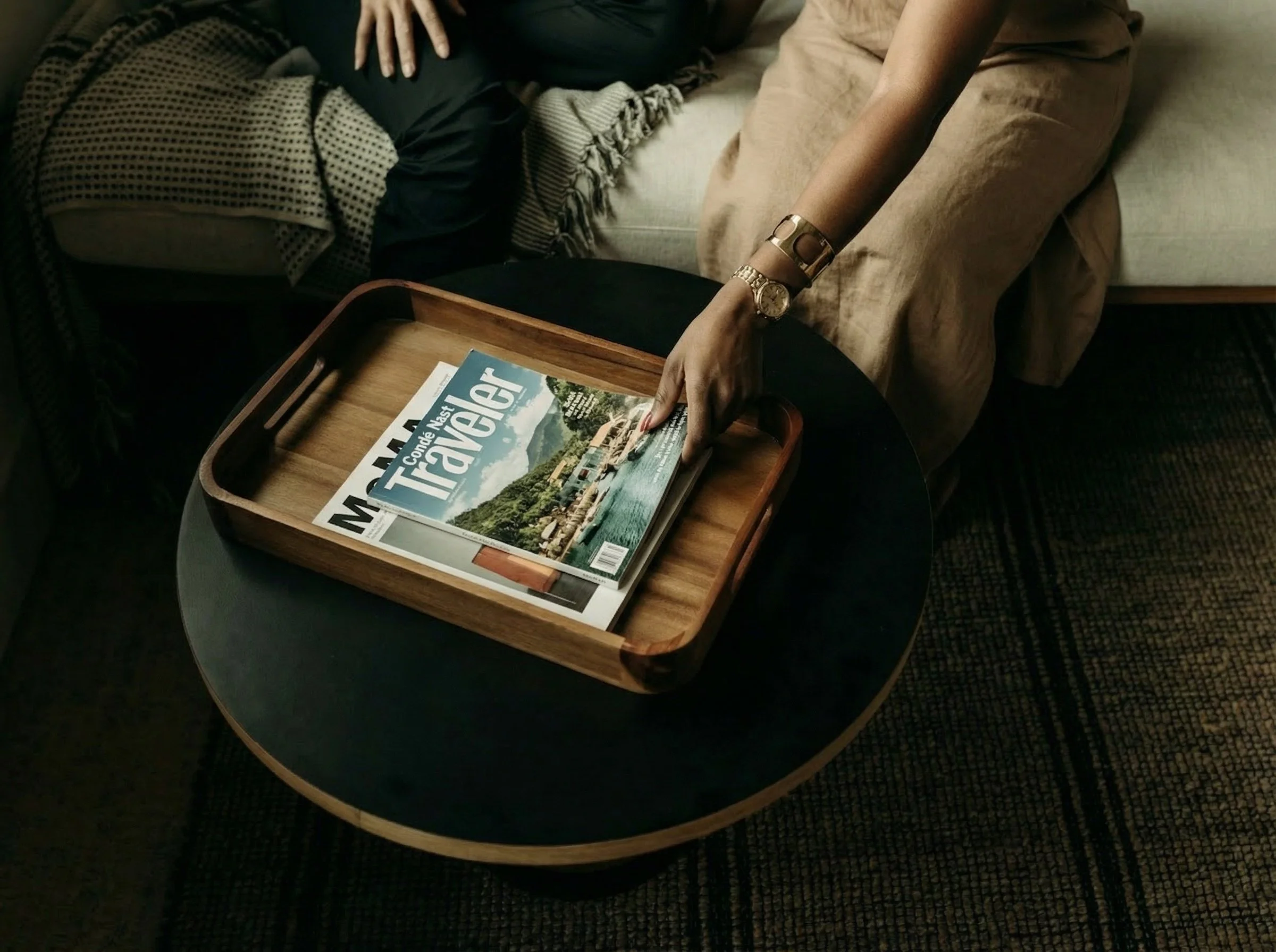 A person is sitting on a couch with a blanket, reaching over a small dark-colored table to pick up a travel magazine from a wooden tray. The magazine has a cover photo of a lakeside scene and the title "Travel".