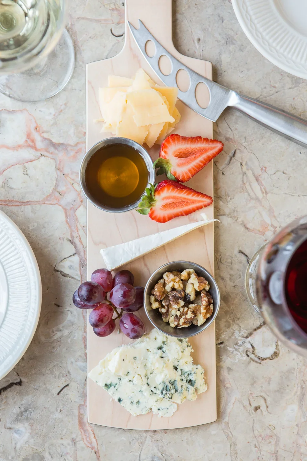 A cheese board with various cheeses, strawberries, grapes, walnuts, honey, and a glass of red wine on a marble table.