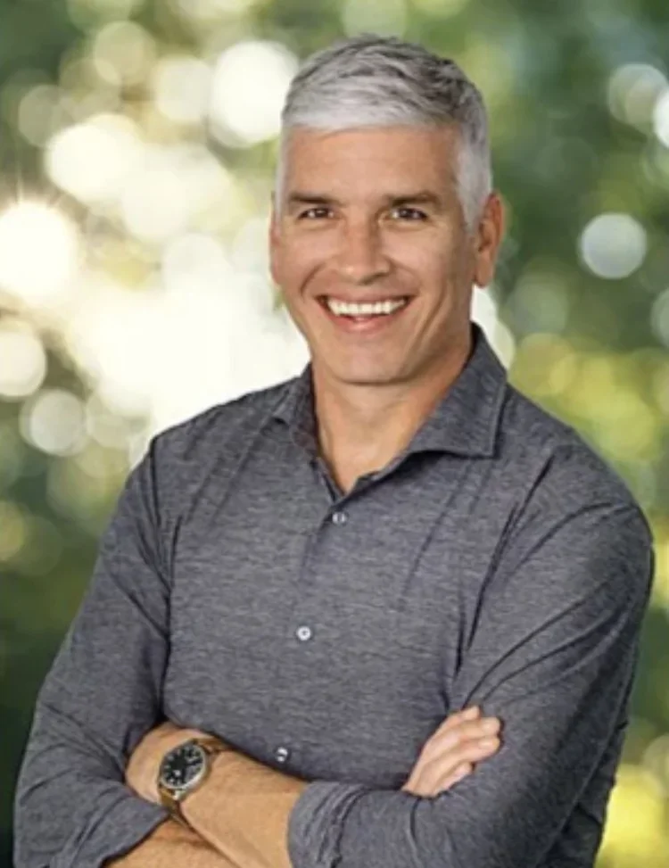 A smiling middle-aged man with short gray hair, wearing a dark gray button-up shirt and a watch, standing outdoors with a blurred green background.