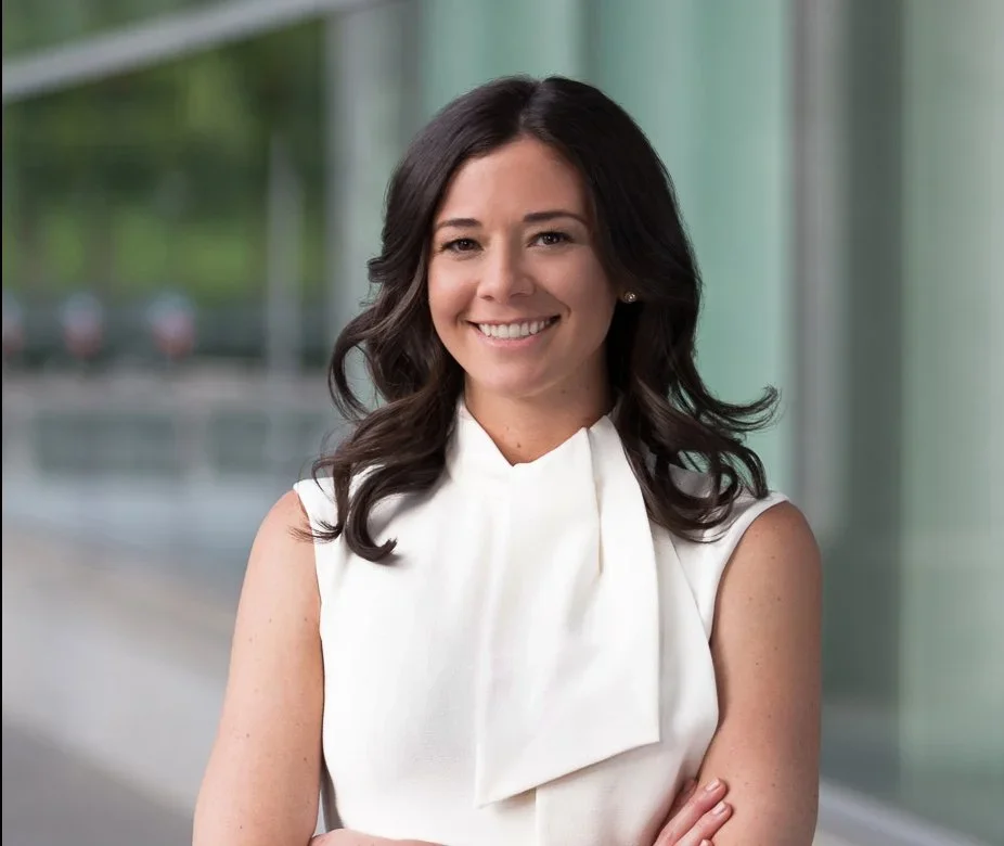 Smiling woman with dark wavy hair in a sleeveless white blouse, standing in front of a modern building with glass windows.