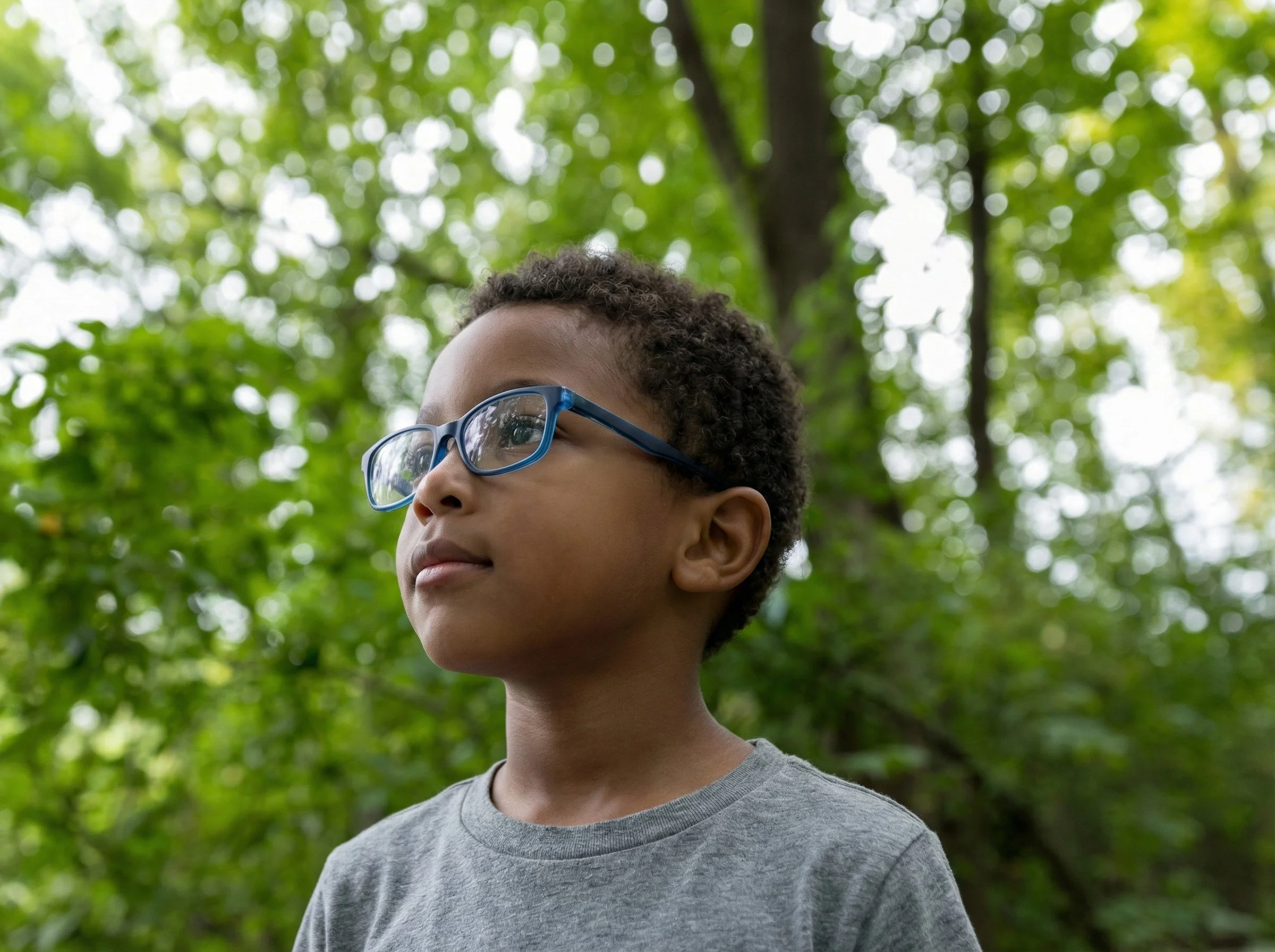 A young boy with curly hair and glasses looking thoughtfully into the distance outdoors surrounded by green foliage.