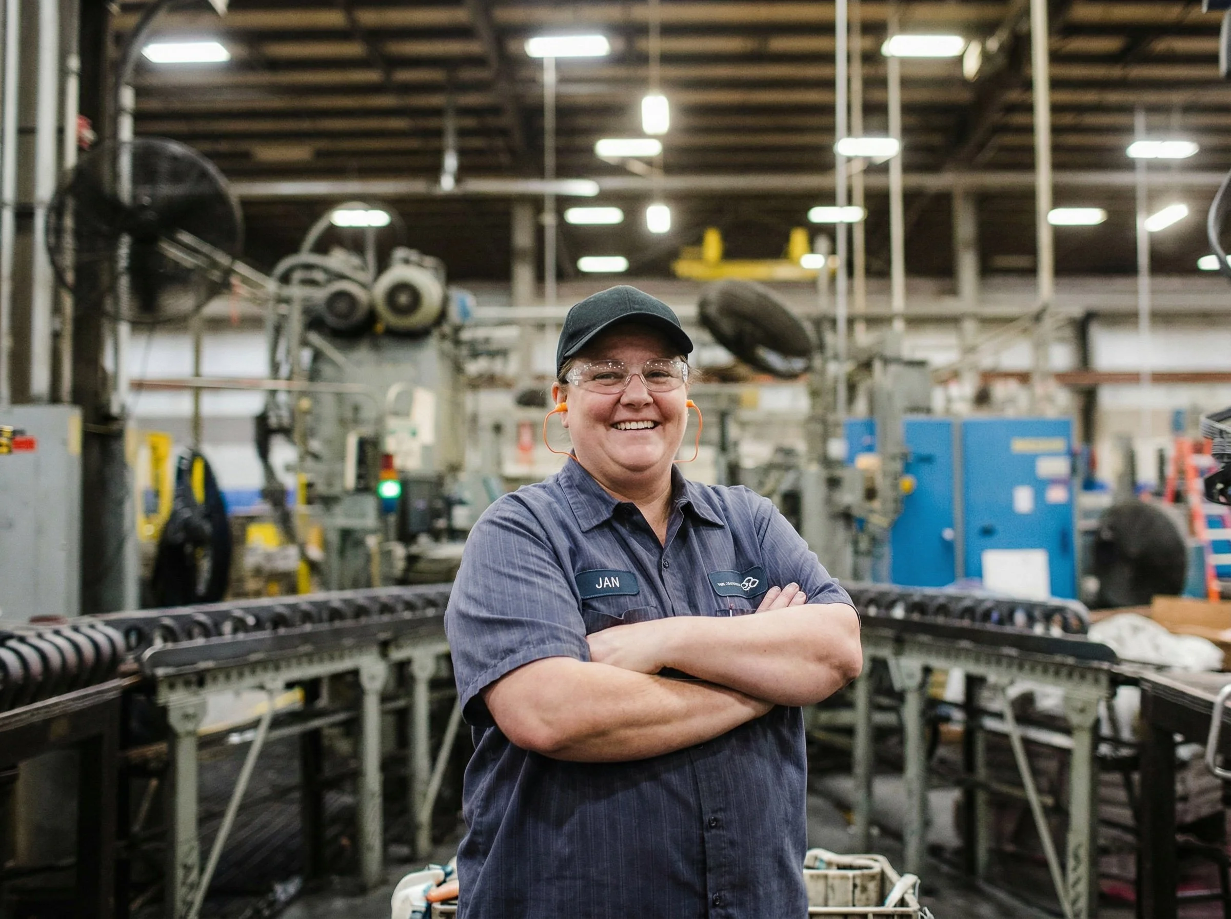 A smiling woman with crossed arms, wearing safety glasses, a cap, and a work uniform, standing in an industrial factory or manufacturing setting with machinery and conveyor belts