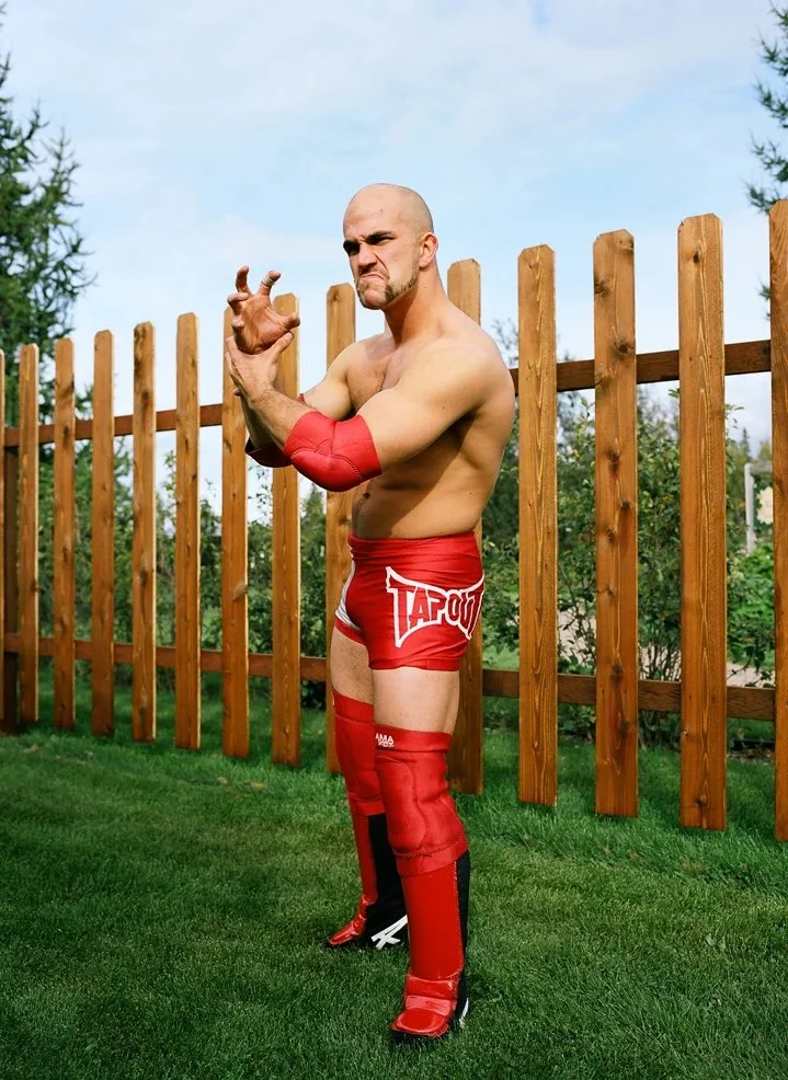 A muscular man in red sports gear posing outdoors in front of a wooden fence, with a stern expression.