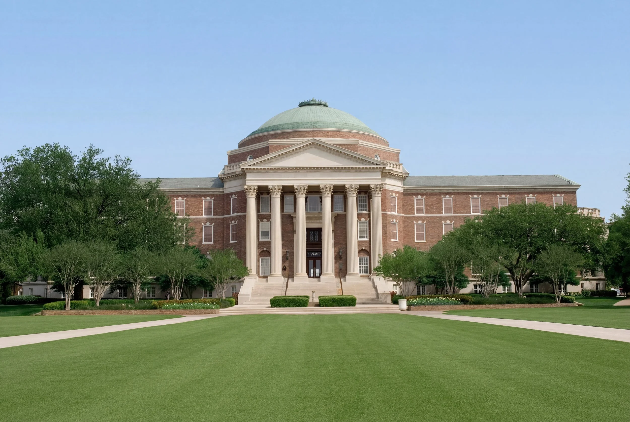 A large, formal government or university building with a domed roof, brick facade, tall columns at the entrance, surrounded by green lawns and trees.
