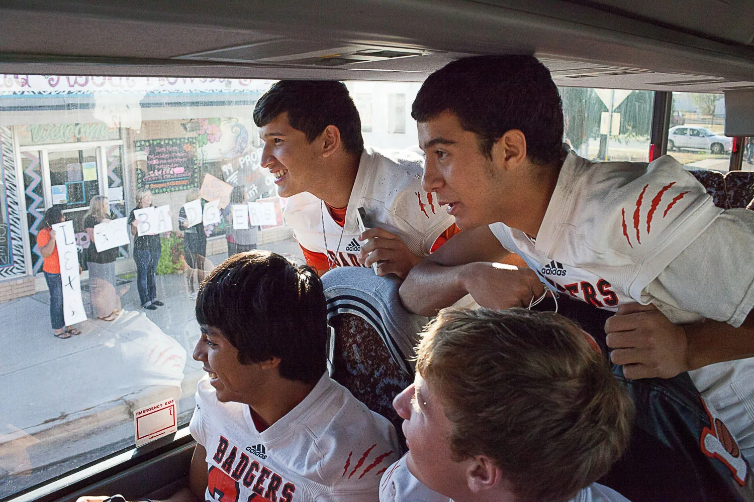Group of young football players wearing white jerseys with red and black accents, leaning inside a bus and looking out the window, smiling and chatting.