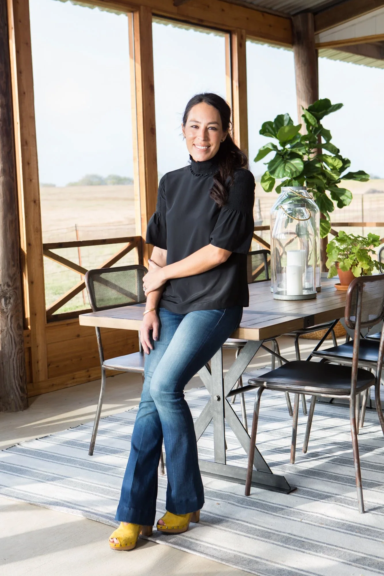 A woman standing indoors near a wooden table and chairs, with a large window and a view of a field outside. She is smiling, wearing a black top, blue jeans, and yellow shoes, and has dark hair.