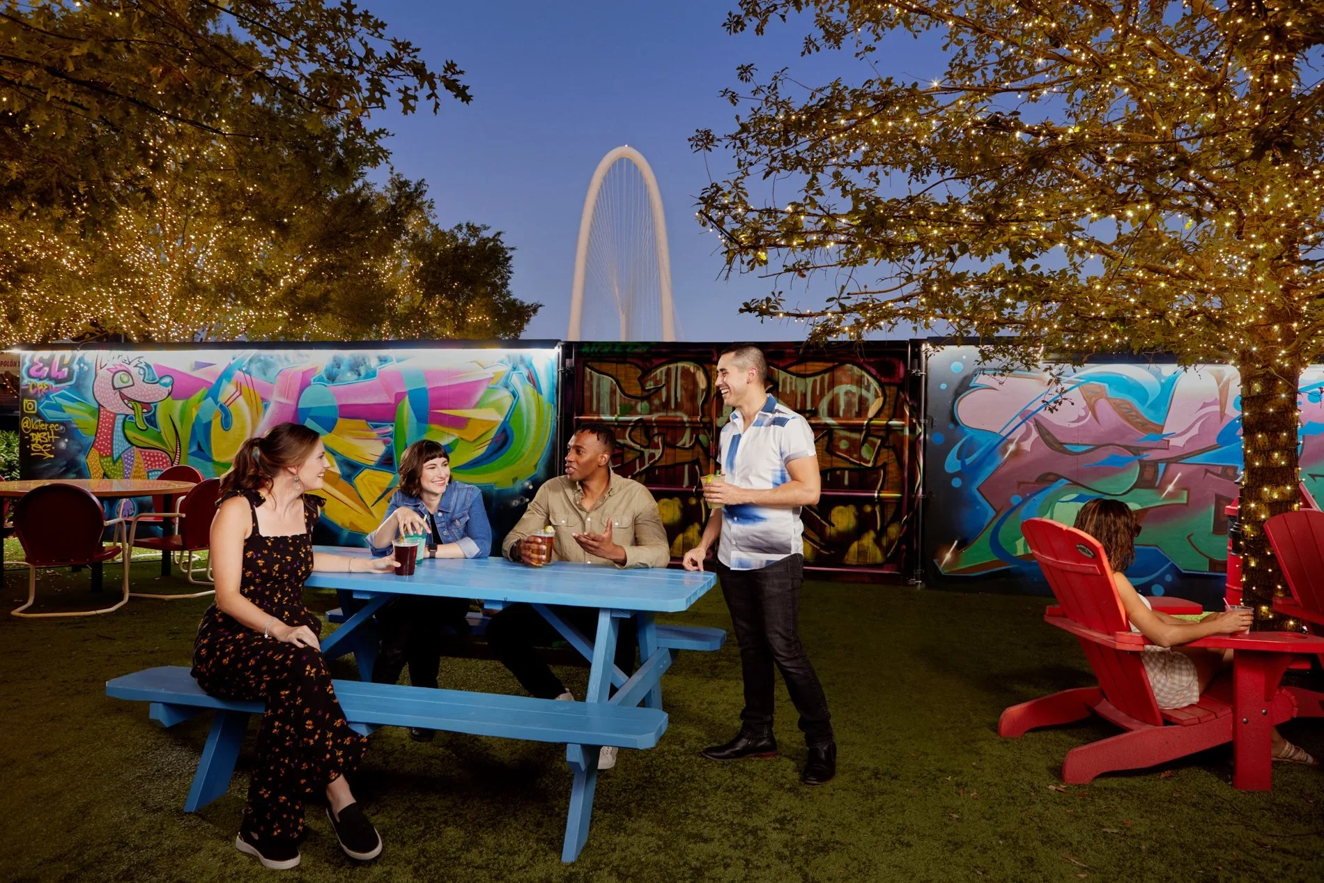 A group of five people socializing outdoors at dusk with colorful graffiti, string lights on trees, and the Ferris wheel in the background. Four are seated around a blue picnic table, and one is standing holding a drink. One person is sitting on a re