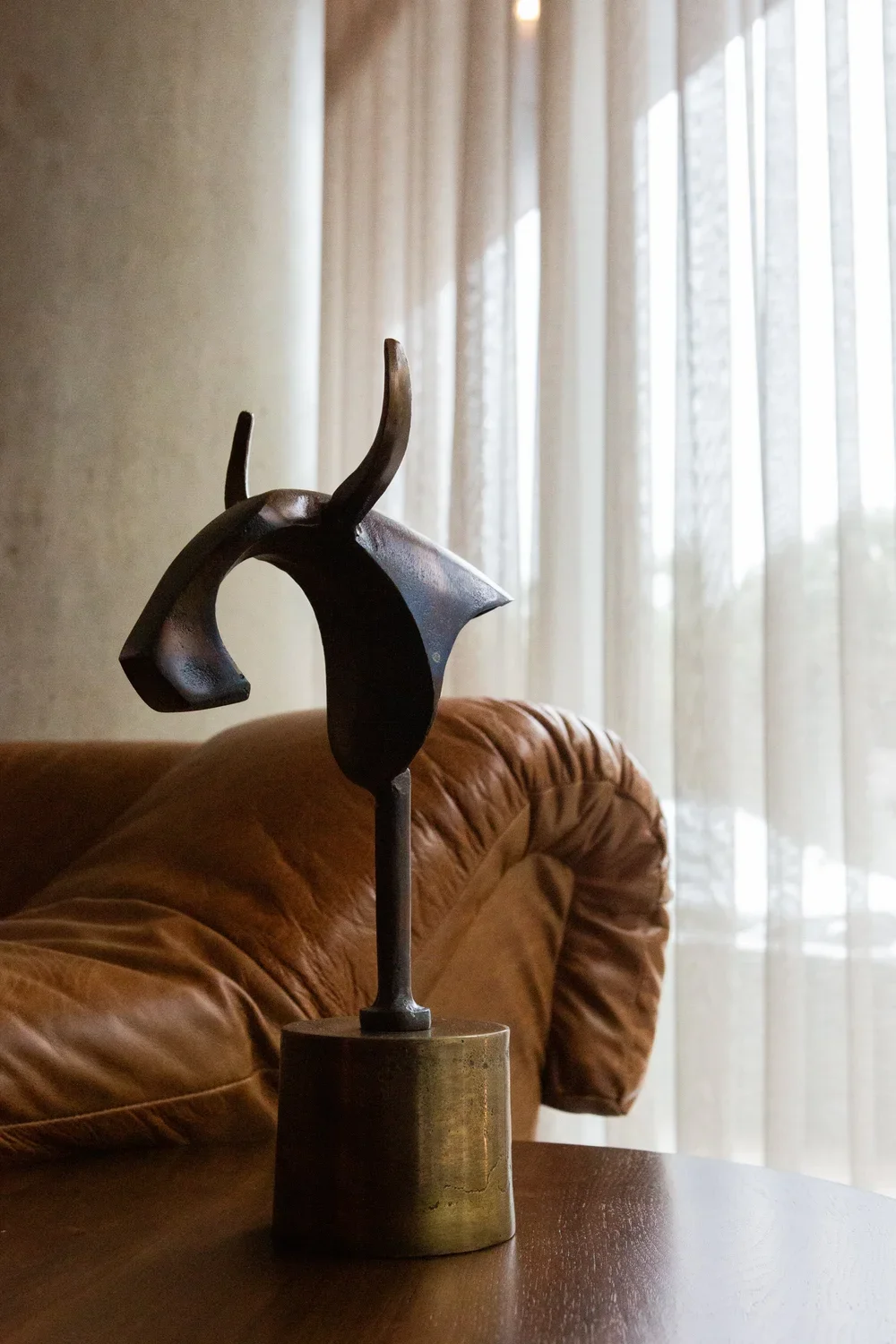 A modern abstract metal sculpture on a gold-colored pedestal, placed on a wooden surface with a leather sofa and sheer curtains in the background.