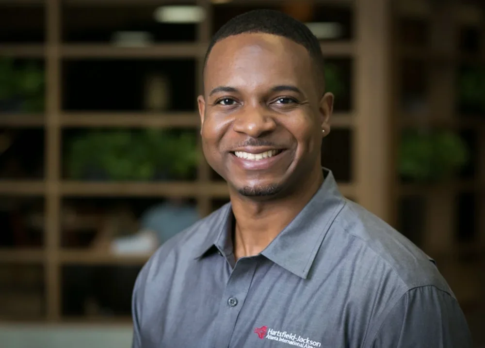 A smiling man with short black hair, wearing a gray collared shirt with the Hartfield Jackson Atlanta International Airport logo, standing in front of a blurred background of wooden shelves and green plants.