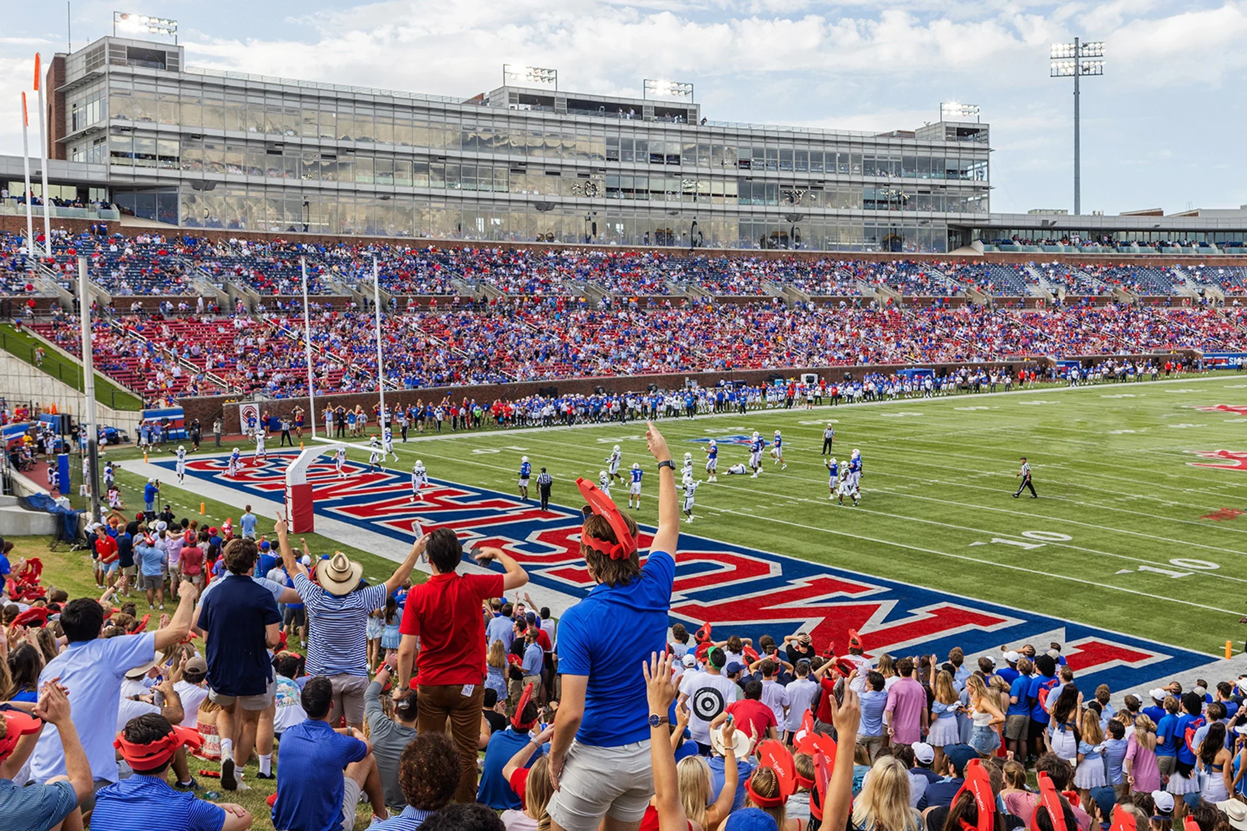 Crowd of spectators watching a football game at a stadium, with fans cheering and some raising their arms, players on the field, and a modern glass-fronted building in the background.