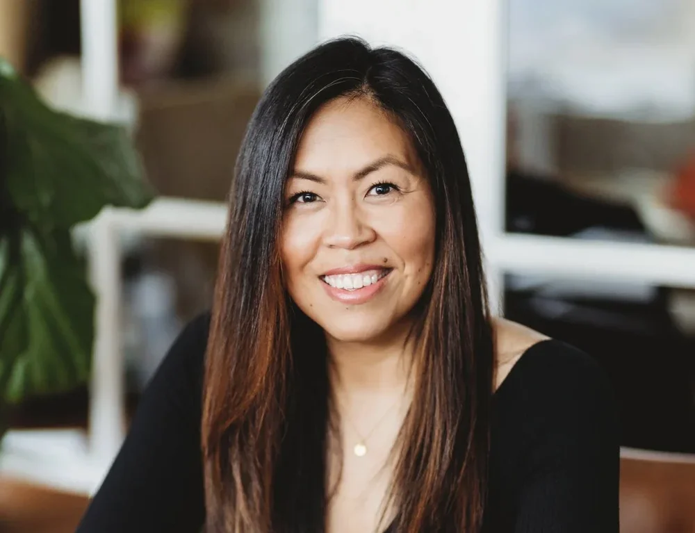 A woman with long dark hair smiling indoors, with a large green plant and blurred background.