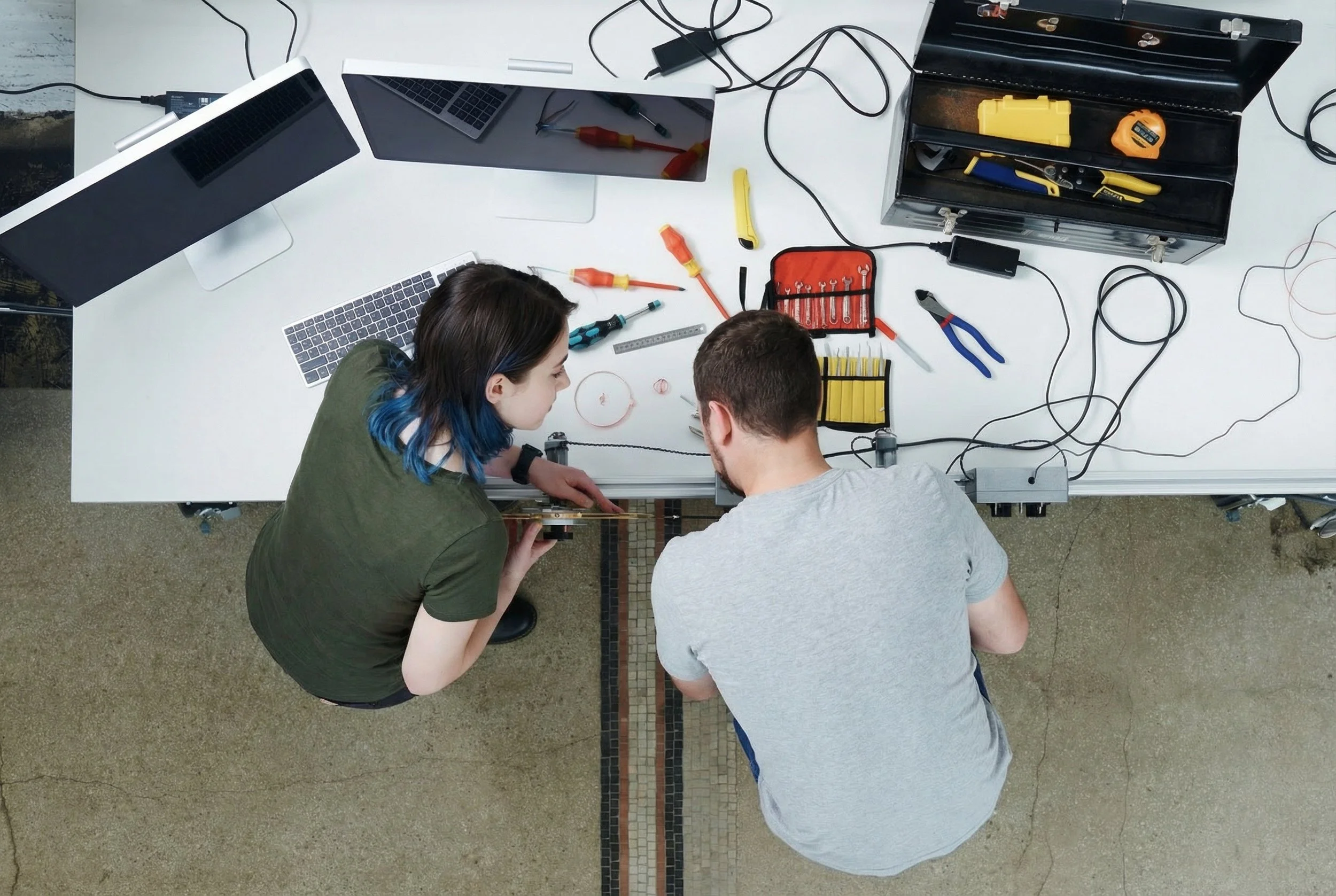 Two people working on a maintenance or engineering project at a white work table filled with tools, wires, and electronic components, viewed from above.
