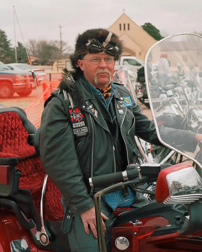 A man wearing a black leather jacket with patches, a fur hat, and glasses, sitting on a motorcycle with a red seat and windshield, in a parking lot with other vehicles and a church building in the background.