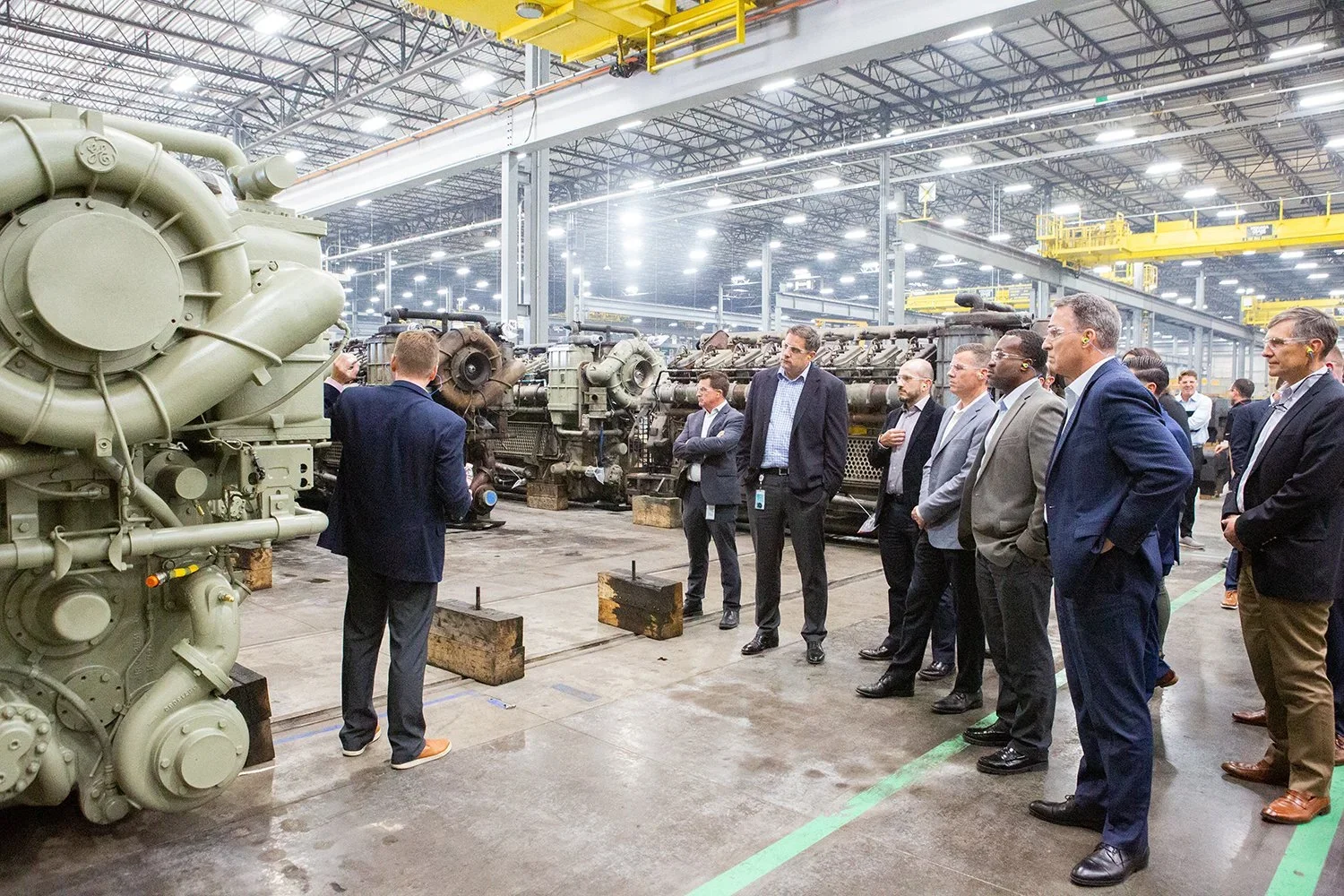 Group of professionals in business attire touring an industrial factory in Plano with large machinery, engines on display.