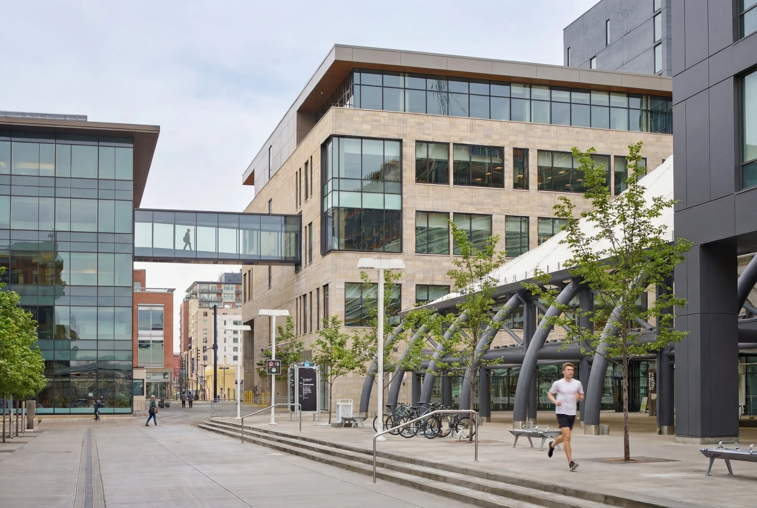 Modern cityscape with office buildings, pedestrian skywalk, and people walking and jogging in the open plaza with trees and benches.