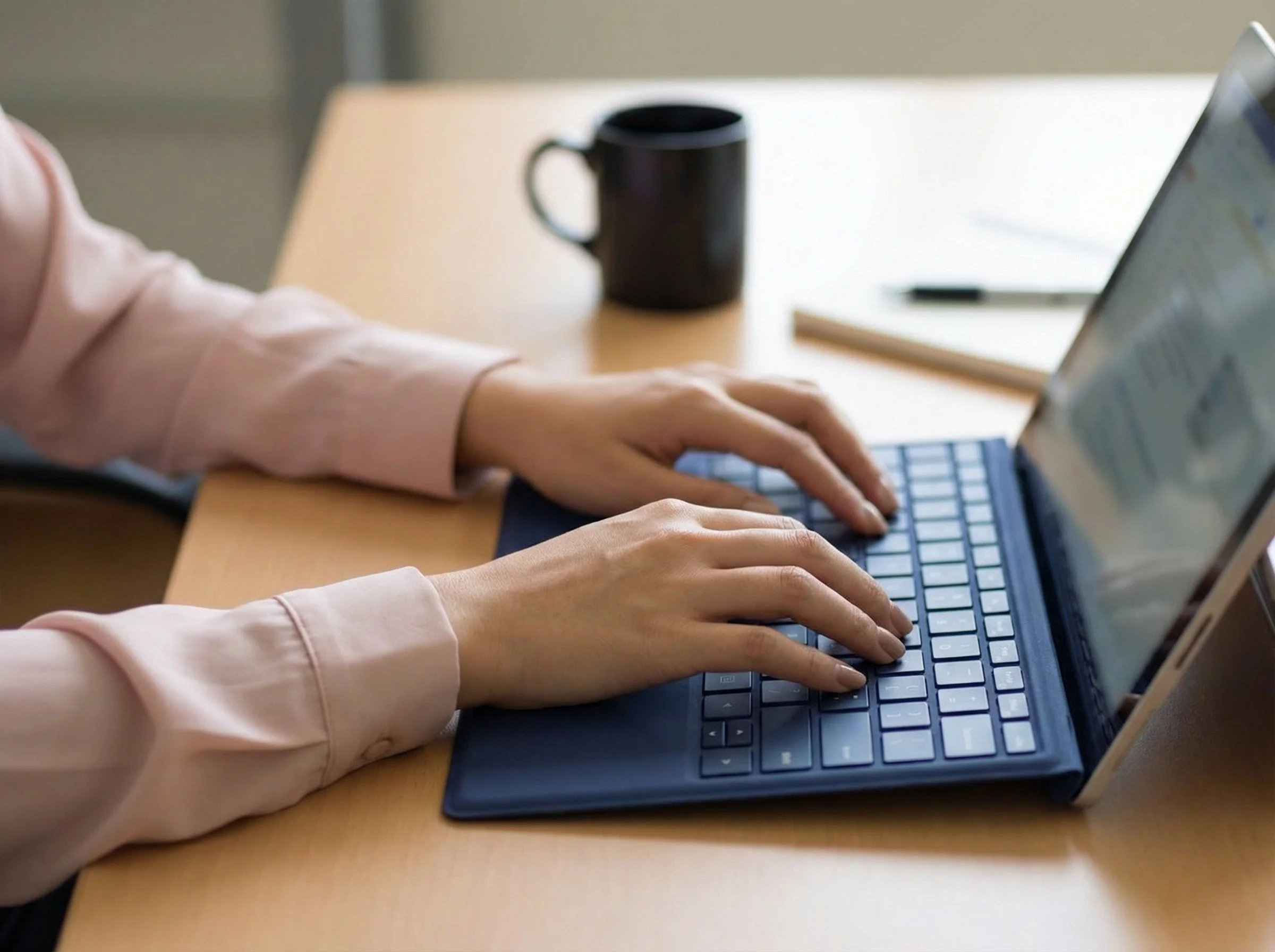 Person typing on a laptop with a black coffee mug and a pen on a wooden table