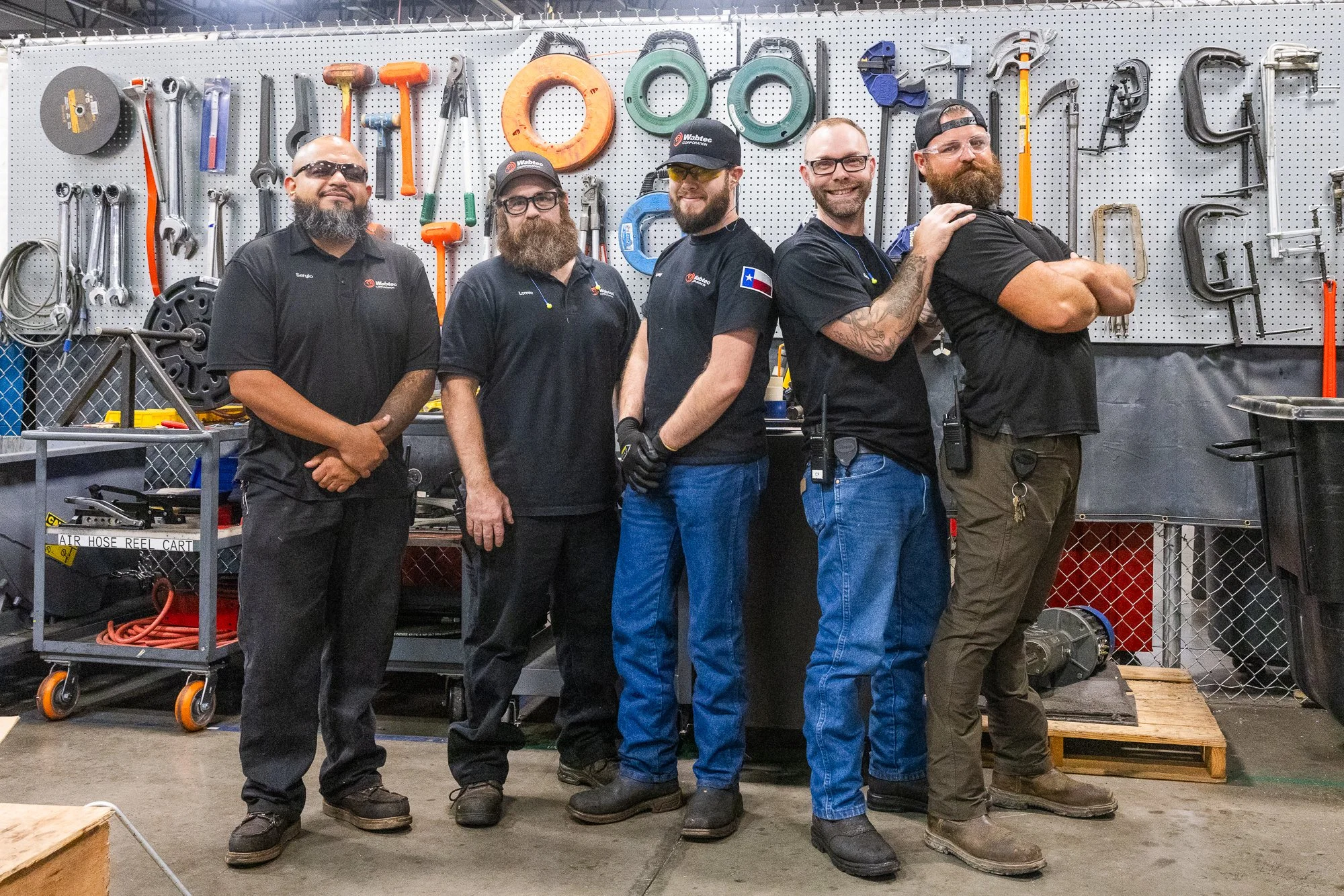 Five men standing in front of a pegboard wall filled with tools, all wearing dark work uniforms, some with gloves and patches. They are smiling and posing for the photo in a workshop.